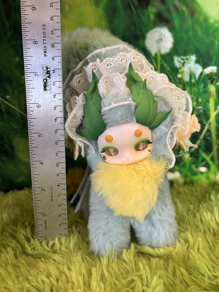 Tutulong Herbalist plush doll standing on a green grass mat next to a vertical metal ruler showing a height of around 8 inches.