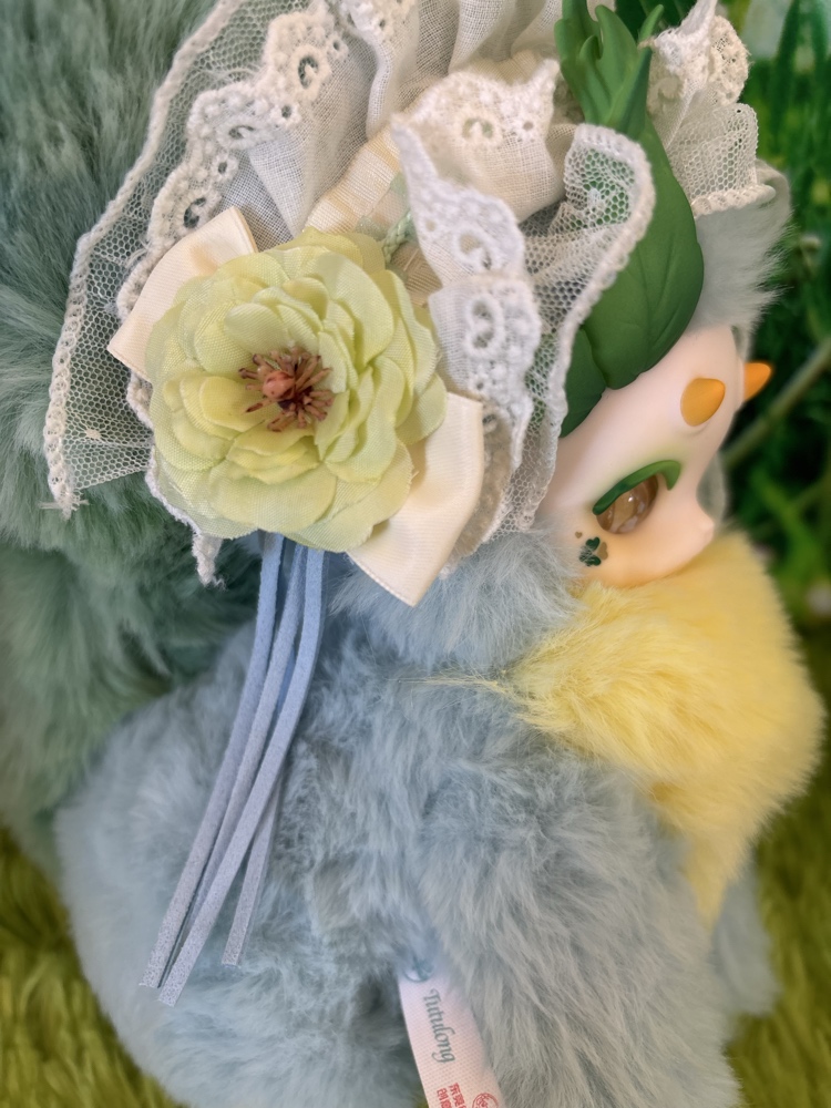 Profile close up of Tutulong Herbalist plush focusing on a chartreuse fabric flower with an orange center and blue hanging ribbons.